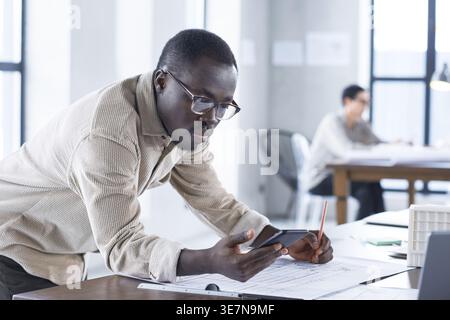 Black male architect wearing eyeglasses using smartphone drawing blueprint while standing and leaning on table near caucasian female colleague in o... Stock Photo