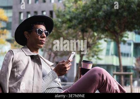 Hispanic man wearing fedora hat and sunglasses holding takeaway coffee and using smartphone sitting with crossed legs on bench in city during daytime Stock Photo