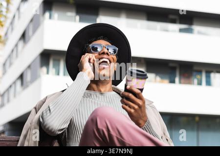 Hispanic man wearing fedora hat and sunglasses holding takeaway coffee and having phone call laughing sitting with crossed legs on bench in city du... Stock Photo