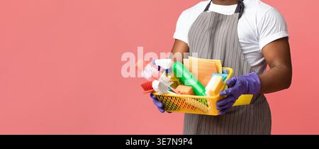 Cropped image of Black man with beard wearing apron and rubber gloves carrying basket of cleaning supplies standing against pink background in studio Stock Photo
