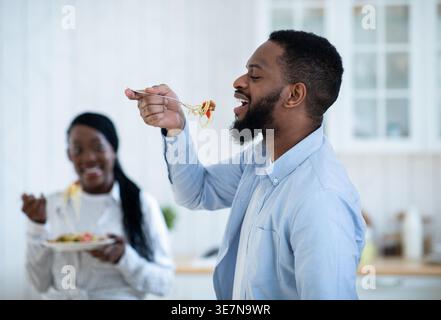 Black man eating spaghetti using fork standing in kitchen near wife at home Stock Photo