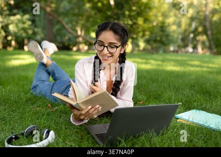Indian female student wearing eyeglasses reading book while lying on grass with laptop and notepads in campus park during daytime Stock Photo