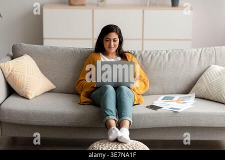 Smiling Middle Eastern woman wearing headsets using laptop sitting on sofa with papers in living room at home Stock Photo