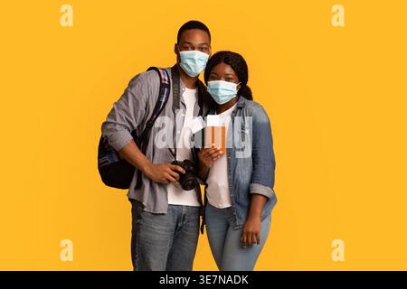 Black couple with face mask carrying backpack and holding passports standing against yellow background in studio Stock Photo