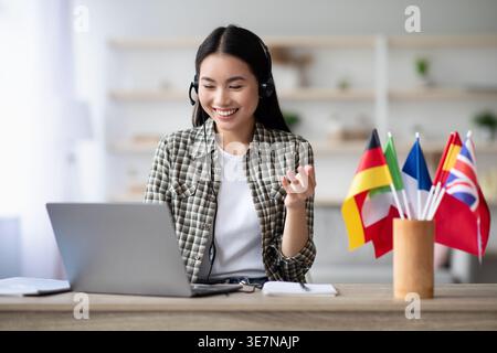 Smiling Asian woman wearing wireless headphones  having video call using laptop while sitting at table with international flags at home Stock Photo
