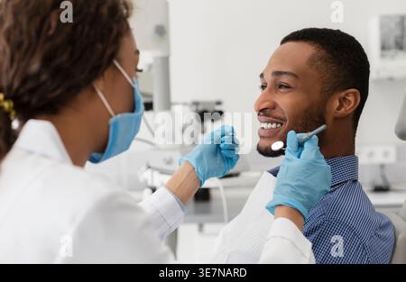 Black male patient getting his teeth checked by female dentist wearing gloves and face mask while sitting on chair in clinic Stock Photo