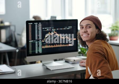 Mixed-race male IT operator looking over shoulder sitting against computer monitor with graphic data on screen near caucasian female colleague in o... Stock Photo