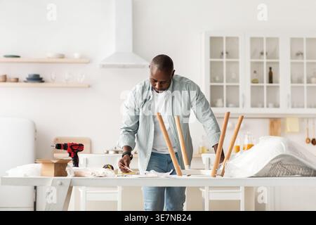 Black man reading instructions on assembling chair standing at table in kitchen at home Stock Photo