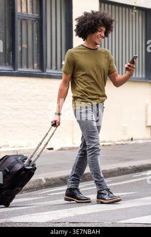 Smiling African American man using smartphone walking with suitcase on crosswalk Stock Photo