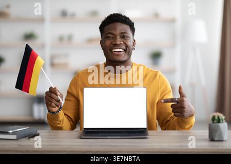 Black man holding flags and pointing finger at laptop screen while sitting at table at home office Stock Photo