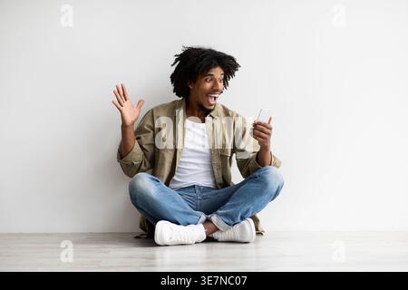 Great News. Surprised Young Black Guy Looking At Smartphone Screen With Excitement While Sitting On Floor Indoors, Emotional African American Man Rais Stock Photo
