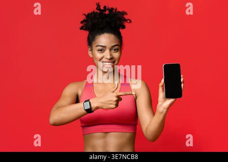 Black woman pointing to smartphone and standing against red background in studio Stock Photo