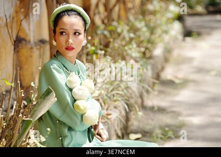 Portrait of attractive young Asian woman in traditional dress and headwear holding lotus flowers and looking away Stock Photo
