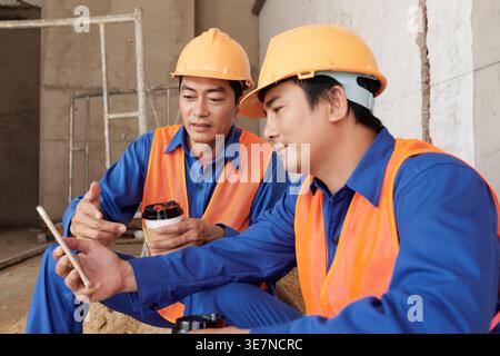 Asian male construction workers with coffee cup sharing smartphone while sitting inside building in construction site Stock Photo