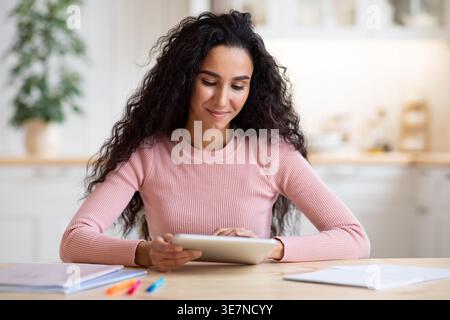 Modern Technologies Concept. Portrait Of Smiling Brunette Woman Using Digital Tablet In Kitchen, Attractive Young Female Sitting At Table With Tab Com Stock Photo