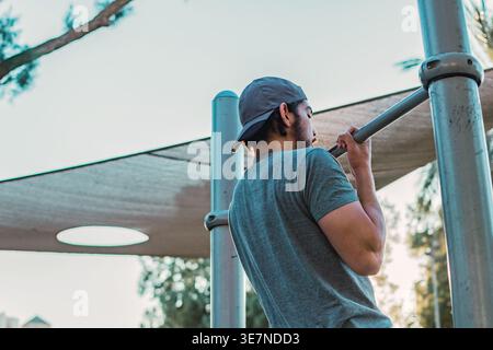 Middle Eastern man with beard wearing cap doing pull-ups in outdoor gym during sunny day Stock Photo