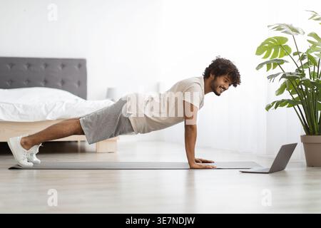 Asian man with curly hair watching fitness video on laptop while planking on yoga mat in bedroom at home Stock Photo