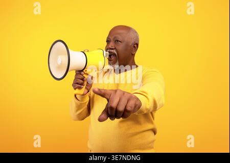 Middle-aged Black man wearing yellow sweater pointing finger away screaming through megaphone while standing against yellow background in studio Stock Photo
