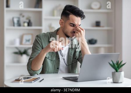 Tired Arab man taking off eyeglasses massaging nose bridge sitting at table with laptop at home Stock Photo