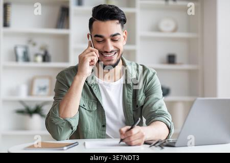 Arab man having phone call writing down notes sitting at table with laptop at home Stock Photo