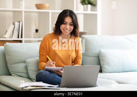 Smiling Hispanic woman taking down notes while working on laptop sitting on bed in bedroom at home Stock Photo