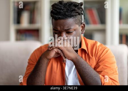 Depressed Black man leaning mouth on clasped hands sitting on sofa in living room at home Stock Photo