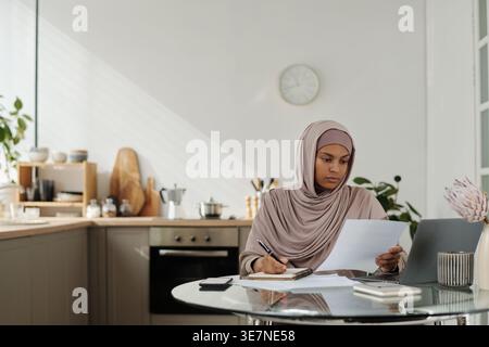 Black female teenager in hijab reading documents and making notes while sitting at table in living room at home Stock Photo