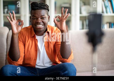 Smiling Black man with braids showing OK hand gestures sitting on sofa in living room at home Stock Photo