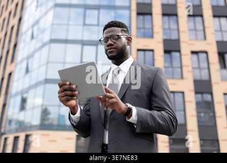 Black businessman in eyeglasses using tablet standing on city street during daytime Stock Photo