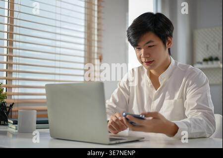 Asian man using smartphone while working on laptop sitting at table at home office during daytime Stock Photo