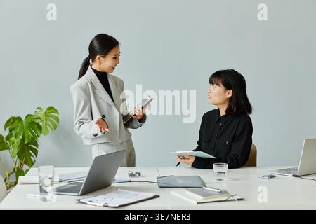 Smiling Asian women discussing business project standing and sitting at table with laptop and glass of water against gray wall in office Stock Photo