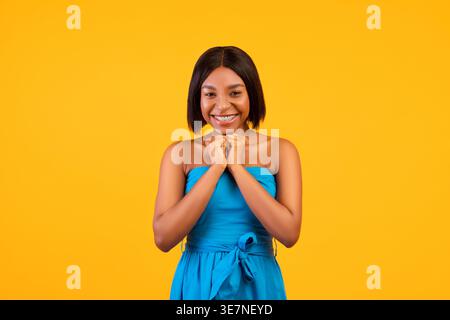 Black woman in blue dress putting hands together standing against orange background in studio Stock Photo