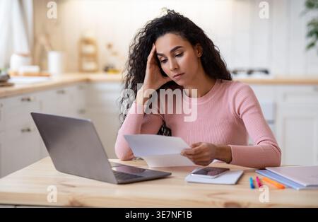 Stressed Female Entrepreneur Working With Papers And Laptop At Home, Concerned Millennial Woman Sitting At Table In Kitchen, Reading Financial Documen Stock Photo