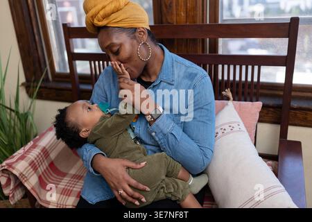 Black mother wearing yellow head wrap and long sleeve denim shirt carrying and playing with her baby boy with pacifier while sitting on wooden sofa... Stock Photo
