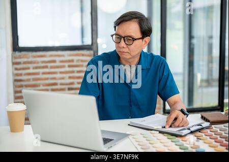 Asian middle-aged male interior designer looking at laptop sitting at the table with materials in office Stock Photo