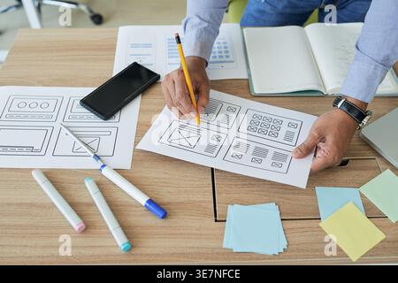 Indian male designer correcting webpage layout document with pencil while leaning on desk with smartphone in office Stock Photo
