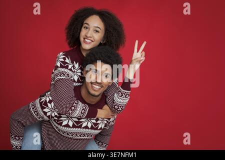 Black man with afro wearing red sweater giving piggyback ride to girlfriend making peace hand sign against red background in studio Stock Photo