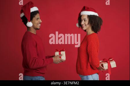 Smiling Black man in red sweater wearing Santa hat giving gift box to girlfriend while standing against red background in studio Stock Photo