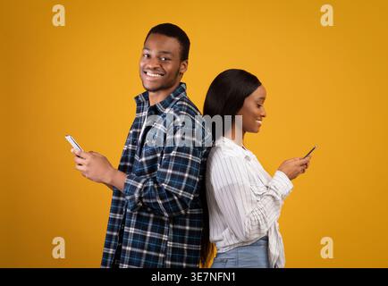 Black couple using smartphones while standing back to back against yellow background in studio Stock Photo