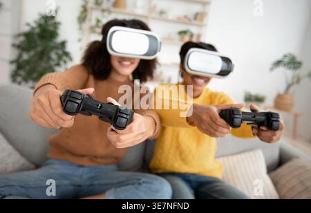 Black female friends wearing VR experiencing virtual reality using game controller having fun while sitting on sofa in living room at home Stock Photo