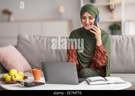 Middle-aged Middle Eastern muslim woman in hijab having phone call and using laptop while sitting at coffee table with calculator and notebook in l... Stock Photo