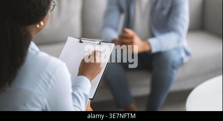 Black female psychotherapist taking notes listening to male patient during therapy session in clinic Stock Photo