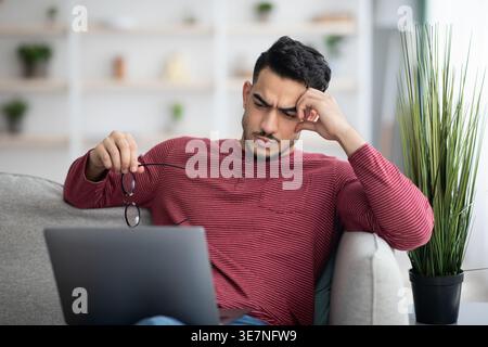 Middle Eastern man holding eyeglasses looking at laptop sitting on couch in living room at home Stock Photo