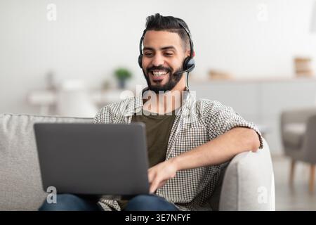 Positive young Arab man wearing headphones, having online meeting on laptop at home. Eastern male student participating in webinar, learning remotely, Stock Photo