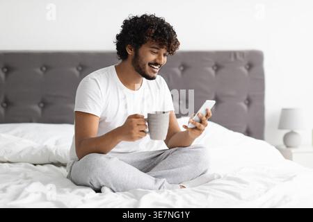 Bearded Indian man with curly hair having video call on smartphone while having coffee sitting on bed in bedroom at home Stock Photo