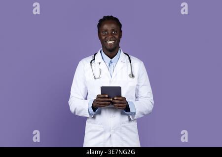Smiling Black male doctor with dreads wearing white suit with stethoscope holding tablet while standing against purple background in studio Stock Photo