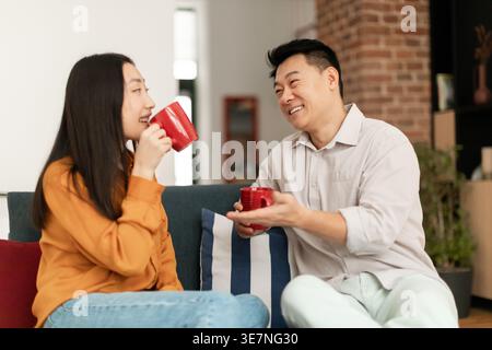 Asian couple having fun drinking coffee and talking while sitting together on sofa in living room at home Stock Photo