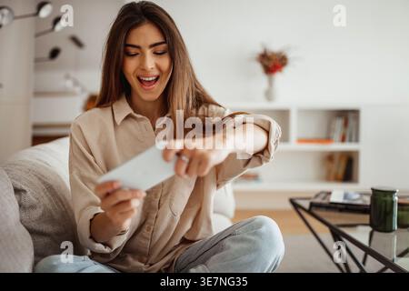 Cheerful Hispanic woman having fun playing games on her smartphone while sitting with crossed legs on sofa in living room at home Stock Photo