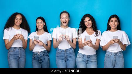 Diverse group of women wearing white shirts and jeans using smartphones standing against blue background in studio Stock Photo