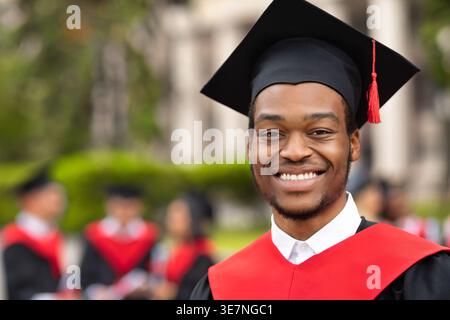 Black male student wearing graduation gown and cap standing near diverse group of students on university campus after graduation ceremony Stock Photo